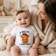 A baby wearing a white bodysuit with a Scottish highland cow graphic and the text 'A WEE BIT SCOTTISH' sits on a cozy cream knit blanket next to a smiling woman in a beige cardigan.