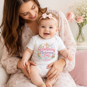 Woman holding a smiling baby wearing a personalised pink christening day bodysuit with a cross and flowers.