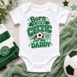 A white short-sleeve baby bodysuit laid flat on a light wood surface, printed with green and black text reading 'Born to support Celtic Just like Daddy' alongside a soccer ball and Celtic crest. A green-and-white striped scarf, green fabric, and a soccer ball are styled around it.