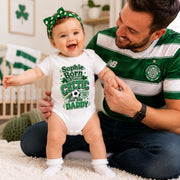 A baby wearing a white bodysuit with a green and white 'Sophie Born to Support Celtic Just like Daddy' graphic stands on a white rug while being held by a man wearing a green and white Celtic FC New Balance jersey.