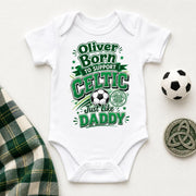 A white short-sleeve baby bodysuit laid flat on a light background, printed with green and gray text reading 'Oliver Born to support CELTIC Just like DADDY' around a soccer ball graphic. A small soccer ball, a Celtic coin, and a plaid fabric strip are placed around it as styling props.