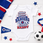 A white short-sleeve baby bodysuit laid flat on a white wood surface, printed with a patriotic red, white, and blue graphic that reads 'Adam Born to Support Rangers Fast like Daddy' with a Rangers Football Club crest. Decorative props include a red, white, and blue striped fabric, a small Rangers flag, stars, ribbon, and a soccer ball.
