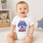 A smiling baby sitting on a white rug wearing a white short-sleeve bodysuit with a red, white, and blue graphic that reads 'Born to Support Rangers Just like Daddy' with a soccer ball and star design.