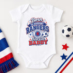 A white short-sleeve baby bodysuit laid flat on a light background, printed with a red, white, and blue graphic that reads 'Born to Support Rangers Just Like Daddy' with a soccer ball and crest. A striped red, white, and blue scarf, a soccer ball, star shapes, and diagonal stripes are styled around it.