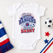 A white short-sleeve baby bodysuit laid flat on a light background, printed with a red, white, and blue graphic that reads 'Born to Support Rangers Just Like Daddy' with a soccer ball and crest. A striped red, white, and blue scarf, a soccer ball, star shapes, and diagonal stripes are styled around it.
