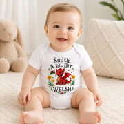 A smiling baby sitting on a cream rug wearing a white short-sleeve bodysuit with a colorful graphic of a red dragon and the text 'Smith A LIL BIT WELSH'.