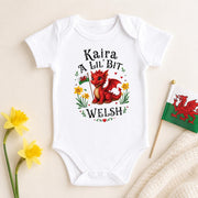 A white short-sleeve baby bodysuit laid flat on a light beige background, printed with a red dragon illustration and the text 'Kaira A Lil' Bit Welsh.' Yellow flowers and a small Welsh flag are placed around it as styling props.