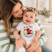 A smiling baby wearing a white bodysuit with a red and green fox graphic and the text 'A Lil' Bit Welsh' sits on a woman's lap. The woman wears a green and white striped long-sleeve shirt, and the baby also wears a matching red, green, and white floral bow headband.