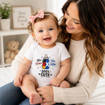 A baby wearing a white short-sleeve bodysuit with a Scottish baby bottle graphic and the text '100% CUTE' sits on a woman's lap. The bodysuit features a baby bottle illustration with a blue and white saltire flag design and '50% Scottish' text.