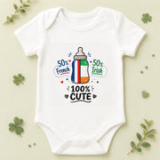 A white short-sleeve baby bodysuit laid flat on a light beige background with decorative green leaves around it. The bodysuit features a colorful graphic of a baby bottle with the Irish flag colors and the text '50% French 50% Irish 100% CUTE'.
