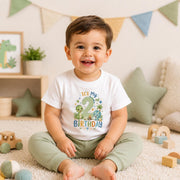 A smiling toddler sits on a cream carpet in a playroom, wearing a white short-sleeve t-shirt with a colorful dinosaur-themed 'It's My 2nd Birthday' graphic and sage green pants.