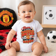 Smiling baby wearing a white “Born to support Manchester United just like Daddy” bodysuit with red and gold lettering.