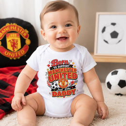 Smiling baby wearing a white “Born to support Manchester United just like Daddy” bodysuit with red and gold lettering.