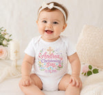 Smiling baby wearing a white personalised pink christening day bodysuit with a cross, flowers and matching headband.