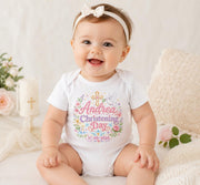 Smiling baby wearing a white personalised pink christening day bodysuit with a cross, flowers and matching headband.