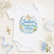White personalised christening day baby bodysuit with blue lettering, name and date, cross and floral decorations.
