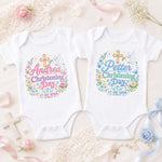 Two white personalised christening bodysuits with pink and blue lettering, names, dates, crosses and flowers.