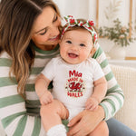 A smiling baby wearing a white short-sleeve bodysuit with a red and green 'Mia Made in Wales' graphic sits on a woman's lap. The woman wears a green and white striped long-sleeve top and a red and green floral bow headband.