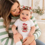 A smiling baby wearing a white short-sleeve bodysuit with a red and green 'Mia Made in Wales' graphic sits on a woman's lap. The woman wears a green and white striped long-sleeve top and a red and green floral bow headband.