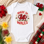 A white short-sleeve baby bodysuit laid flat on a beige fabric background, featuring a circular 'Made in Wales' graphic with a red dragon and small Welsh flags. The bodysuit is styled with a red plush dragon, yellow daffodils, a crocheted textile, a small Welsh flag, and a red-and-green tartan ribbon.