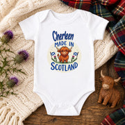 A white short-sleeve baby bodysuit laid flat on a wooden surface, surrounded by a cream knit blanket, purple thistles, a plaid scarf, and a small Highland cow figurine. The bodysuit has a circular graphic that reads 'Cherleen Made in Scotland' with a cartoon Highland cow and Scottish flags.