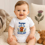 A smiling baby sitting on a cream rug wearing a white short-sleeve bodysuit with a colorful 'Mcgragor MADE IN SCOTLAND' graphic featuring a Highland cow and thistle. A plush Highland cow toy sits to the right, with a decorative pillow and soft home decor in the background.