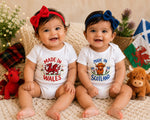 Two smiling babies sitting side by side on a cream knit blanket, wearing white bodysuits with Welsh and Scottish themed graphics. The baby on the left wears a red bow headband and has a white bodysuit printed with 'MADE IN WALES' and a red dragon.