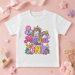 A white short-sleeve kids' t-shirt with a colorful 'Maria Turns One' unicorn graphic, styled in a flat lay on a pink background with decorative props.