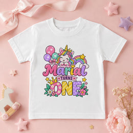 A white short-sleeve kids' t-shirt with a colorful 'Maria Turns One' unicorn graphic, styled in a flat lay on a pink background with decorative props.
