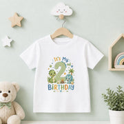 A white short-sleeve children's t-shirt hanging on a wooden hanger against a pale green wall. The shirt features a colorful dinosaur-themed 'It's My 2nd Birthday' graphic.