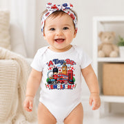 A smiling baby wearing a white short-sleeve bodysuit with a colorful London-themed graphic print and a matching red, white, and blue patterned headband, standing in a bright indoor room.