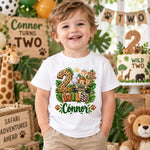 A smiling young boy stands in a decorated safari-themed party setting wearing a white custom graphic T-shirt. The shirt features a colorful '2 Wild Connor' design with safari animals and a jeep, surrounded by green foliage and paw prints.