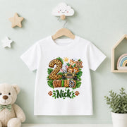 A white kids' t-shirt on a wooden hanger, decorated with a colorful '2nd Birthday Wishes Nick' graphic featuring safari animals and jungle motifs. The shirt is displayed on a light green wall decorated with a teddy bear, wooden stars, and a small plant.