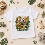 A white kids' t-shirt with a colorful safari-themed '2nd Birthday William' graphic, displayed in a styled flat-lay on a beige background surrounded by toys and leaves.