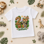 A white kids' t-shirt with a colorful safari-themed '2nd Birthday William' graphic, displayed in a styled flat-lay on a beige background surrounded by toys and leaves.