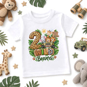 A white short-sleeve kids' t-shirt with a colorful safari-themed graphic reading '2 Wild Beavers' with cartoon animals and a jeep, styled in a flat lay with jungle props.