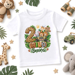 A white short-sleeve kids' t-shirt with a colorful safari-themed graphic reading '2 Wild Beavers' with cartoon animals and a jeep, styled in a flat lay with jungle props.