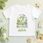 A white short-sleeve baby t-shirt with a colorful dinosaur-themed 2nd birthday graphic, laid flat on a light mint green surface surrounded by toys including a plush dinosaur, wooden stars, and a rainbow.