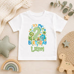 A white short-sleeve baby t-shirt with a colorful dinosaur-themed second birthday graphic reading 'It's My 2 Birthday Liam', styled in a flat-lay on a light wood surface with toys and home decor.