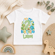 A white short-sleeve baby t-shirt with a colorful dinosaur-themed second birthday graphic reading 'It's My 2 Birthday Liam', styled in a flat-lay on a light wood surface with toys and home decor.