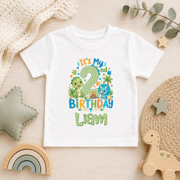 A white short-sleeve baby t-shirt with a colorful dinosaur-themed second birthday graphic reading 'It's My 2 Birthday Liam', styled in a flat-lay on a light wood surface with toys and home decor.