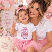 Woman with a smiling child wearing a personalised second birthday T-shirt and pink tutu beside a birthday cake.