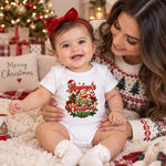 Woman with a smiling baby wearing a personalised first Christmas bodysuit and red bow, beside a Christmas tree.