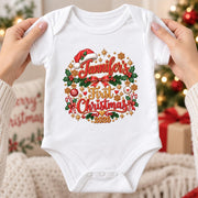 Hands holding a white personalised first Christmas 2026 baby bodysuit with red script, holly and festive decorations.