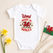 A white short-sleeve baby bodysuit laid flat on a light beige background, printed with a red and cream circular graphic reading 'Richmond Made in Wales' around a red dragon. Yellow flowers and a small Welsh flag are placed as styled props around the bodysuit.