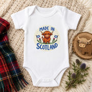 A white short-sleeve baby bodysuit laid flat on a cream knit blanket, featuring a circular 'Made in Scotland' graphic with a Highland cow and Scottish flags. A plaid scarf, a wooden Highland cow slice, and thistle stems are styled around it.