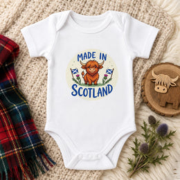 A white short-sleeve baby bodysuit laid flat on a cream knit blanket, featuring a circular 'Made in Scotland' graphic with a Highland cow and Scottish flags. A plaid scarf, a wooden Highland cow slice, and thistle stems are styled around it.