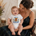 A baby wearing a white short-sleeve bodysuit with a colorful 'Made in Scotland' graphic and lion print, sitting on a woman's lap. The woman is wearing a dark green plaid dress, and the background shows a cozy indoor setting with a candle and dried flowers.
