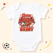 A white short-sleeve baby bodysuit with a colorful graphic print that reads 'Born to Support Arsenal Just Like Daddy' with the Arsenal FC crest and soccer ball motif, displayed on a light beige background with decorative soccer-themed doodles.
