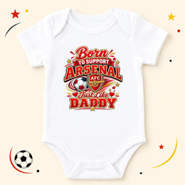 A white short-sleeve baby bodysuit with a colorful graphic print that reads 'Born to Support Arsenal Just Like Daddy' with the Arsenal FC crest and soccer ball motif, displayed on a light beige background with decorative soccer-themed doodles.